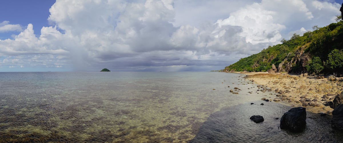 A beach with clear shallow waters, a distant island, and a rocky shoreline with vegetation.