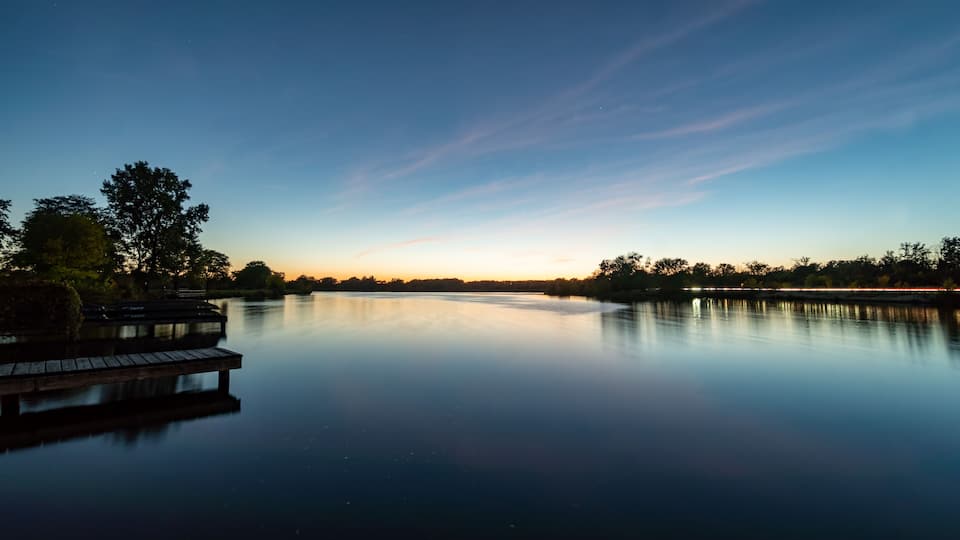A calm lake at dusk with a wooden dock and trees in the background.