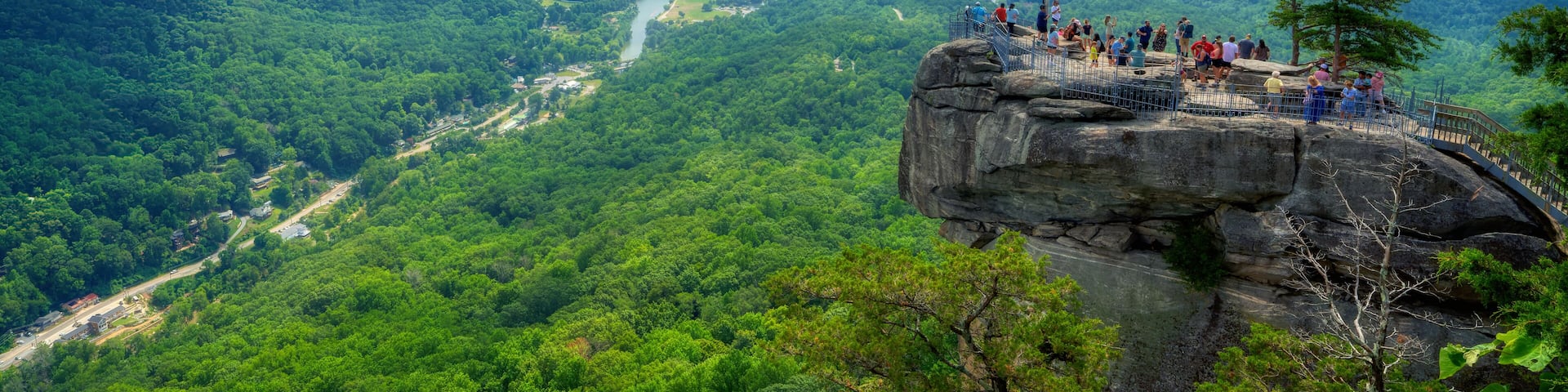 A scenic overlook with an American flag on a rocky cliff overlooking a lush valley and river.
