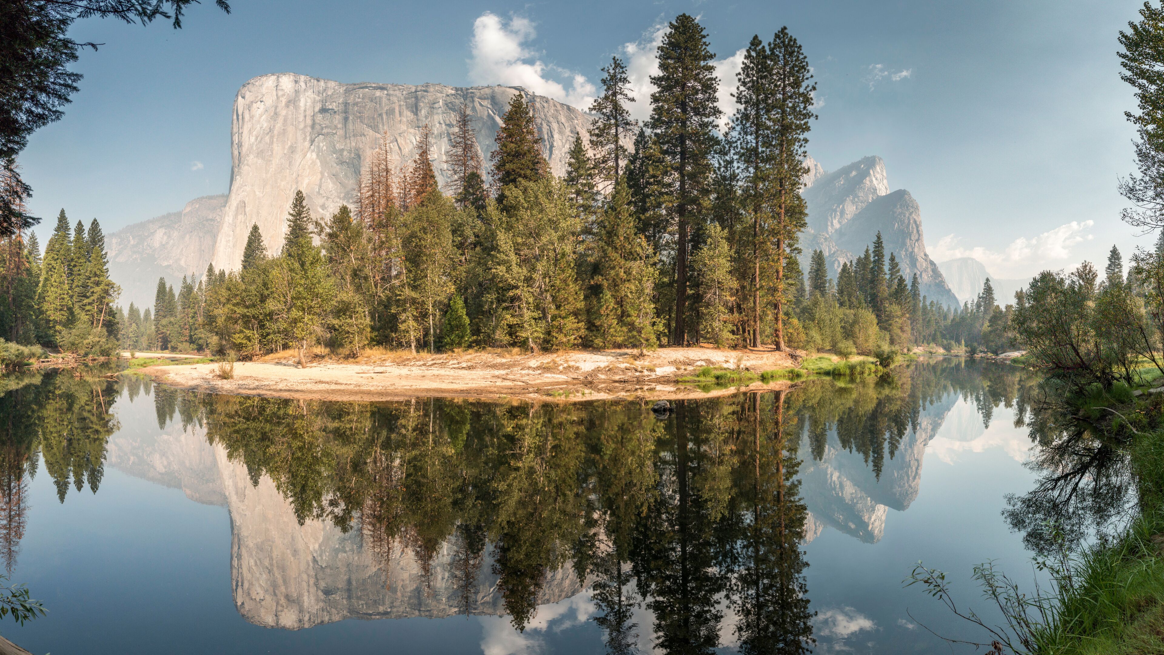 A calm lake reflecting a forest with tall trees and a prominent rock formation in the background.