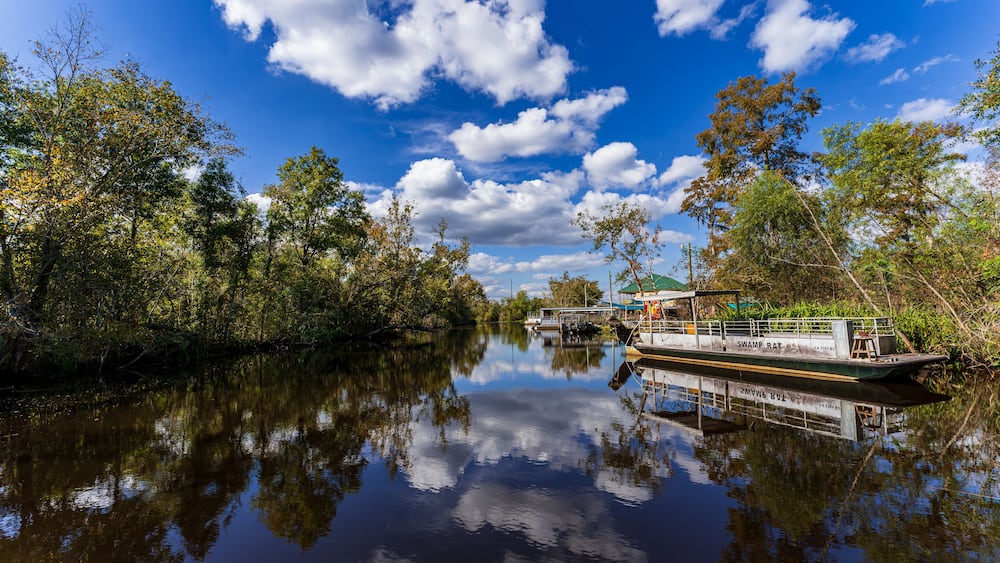 A calm river with a boat docked on the right, surrounded by dense foliage and a clear blue sky with scattered clouds.
