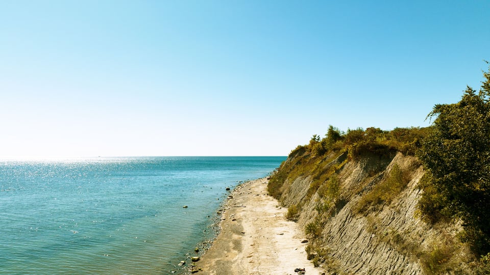A coastal landscape with a clear blue sea, sandy beach, and a steep cliff with sparse vegetation.