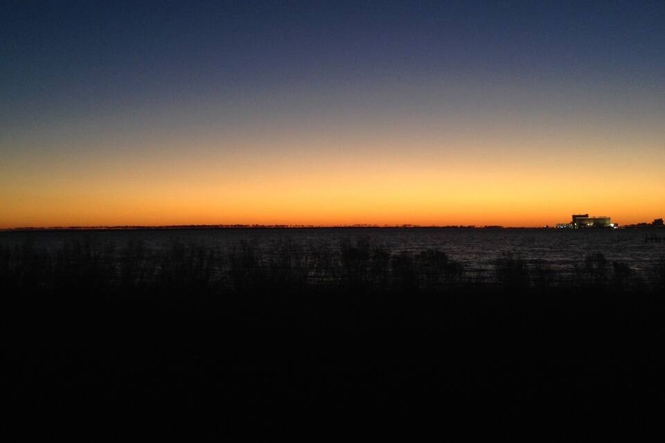 A night sky with a gradient from dark blue to orange near the horizon, and silhouettes of buildings along the coastline.