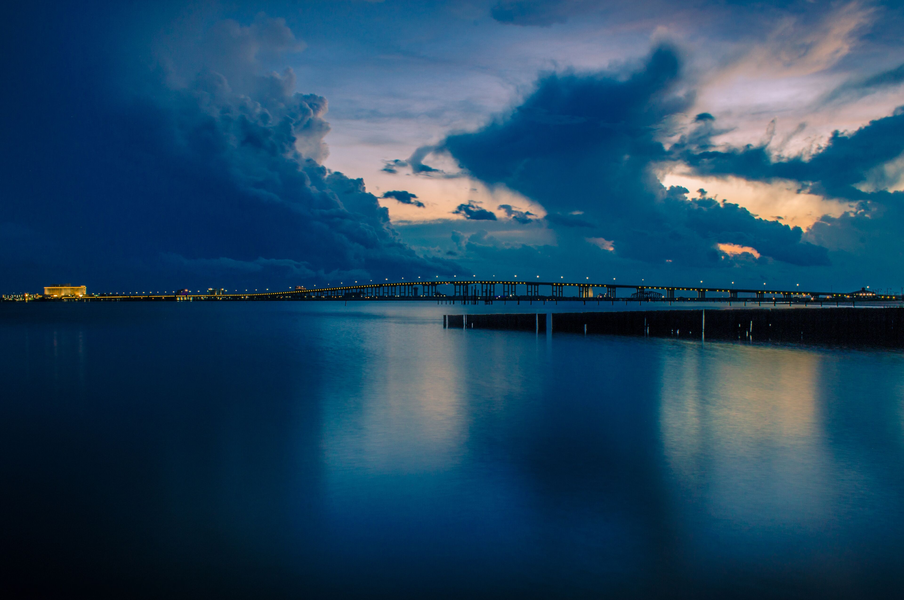 A long pier extending into a calm body of water, with a city skyline in the distance and a dramatic sky with clouds.