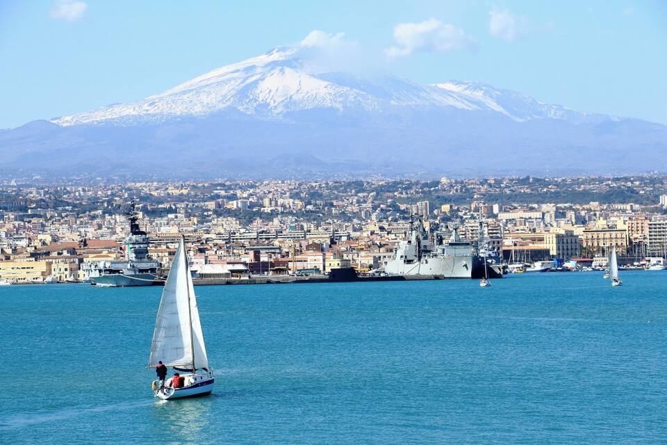 Un velero en un mar tranquilo con un paisaje urbano y una montaña nevada al fondo.
