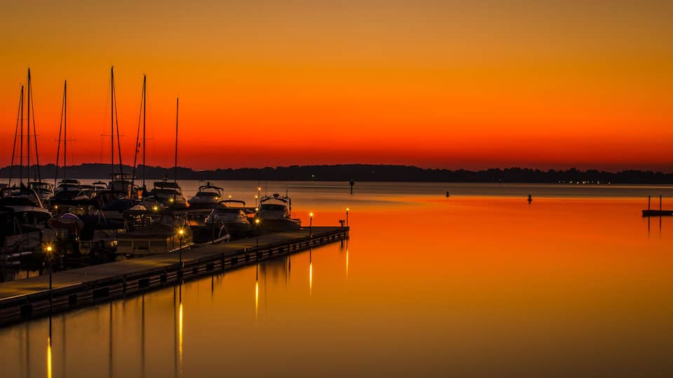 A marina at sunset with boats docked and a calm water surface reflecting the orange sky.