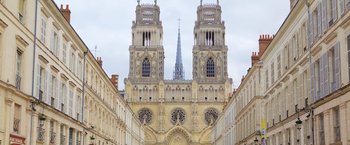 Une imposante cathédrale dotée de deux tours et d’une flèche, bordée de bâtiments richement décorés et entourée d’un espace vert central.