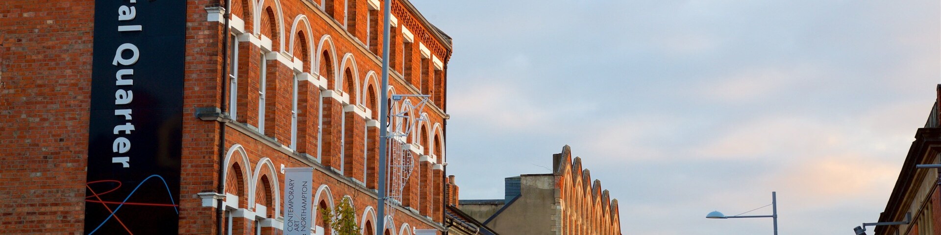 Una calle con edificios de ladrillo rojo, un cartel que indica el Distrito Cultural de Northampton y farolas.
