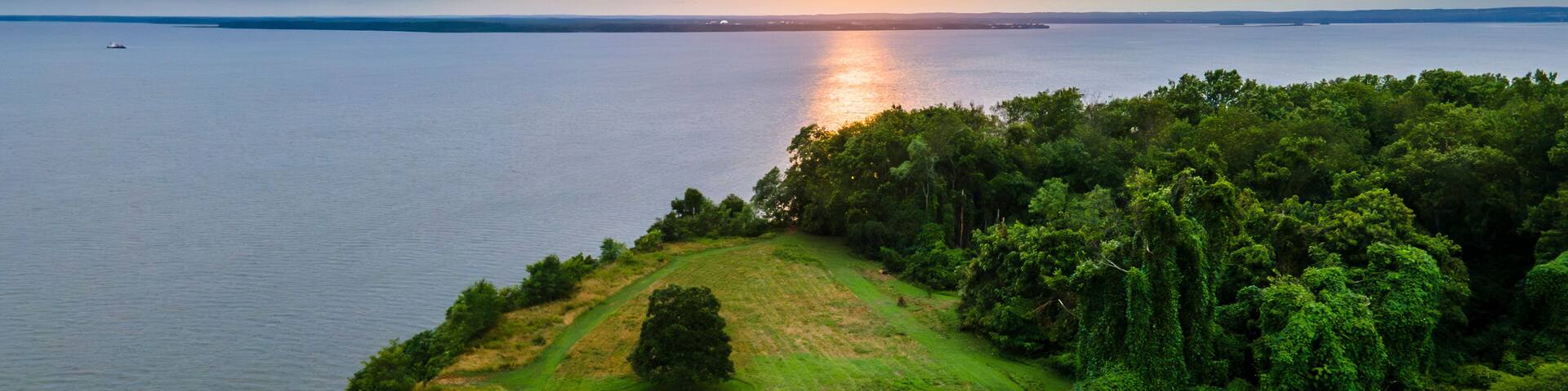 A white lighthouse on a grassy hill overlooking a calm body of water with a sunset in the background.