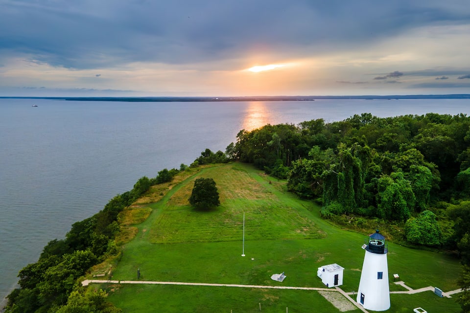 Un phare blanc, perché sur une colline verdoyante, surplombe une étendue d’eau calme, avec un coucher de soleil en arrière-plan.
