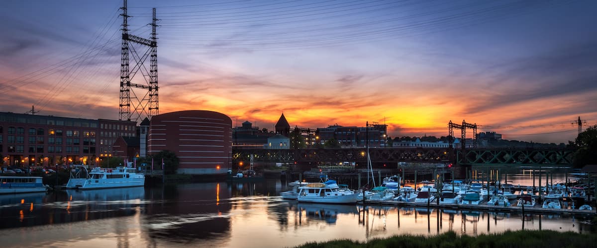 A marina at sunset with boats docked, a bridge, and buildings in the background.