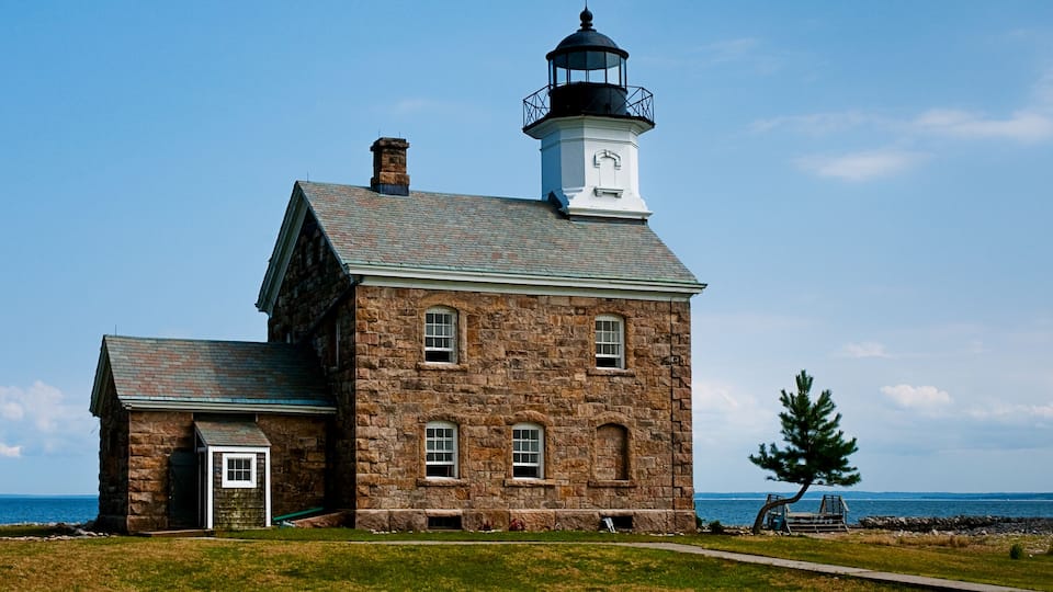 A stone lighthouse with a white tower and black lantern room, situated by the ocean.