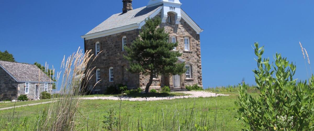 A stone lighthouse with a white tower and black lantern room, surrounded by greenery and a clear blue sky.