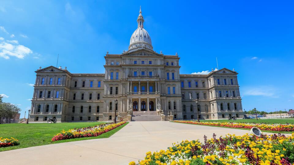 A large neoclassical building with a prominent dome and columns, surrounded by a well-maintained garden with colorful flowers.