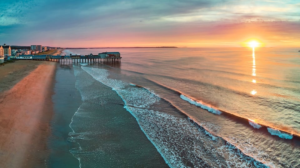 A beach with a pier extending into calm waters, a sunset in the background, and a distant boat on the horizon.