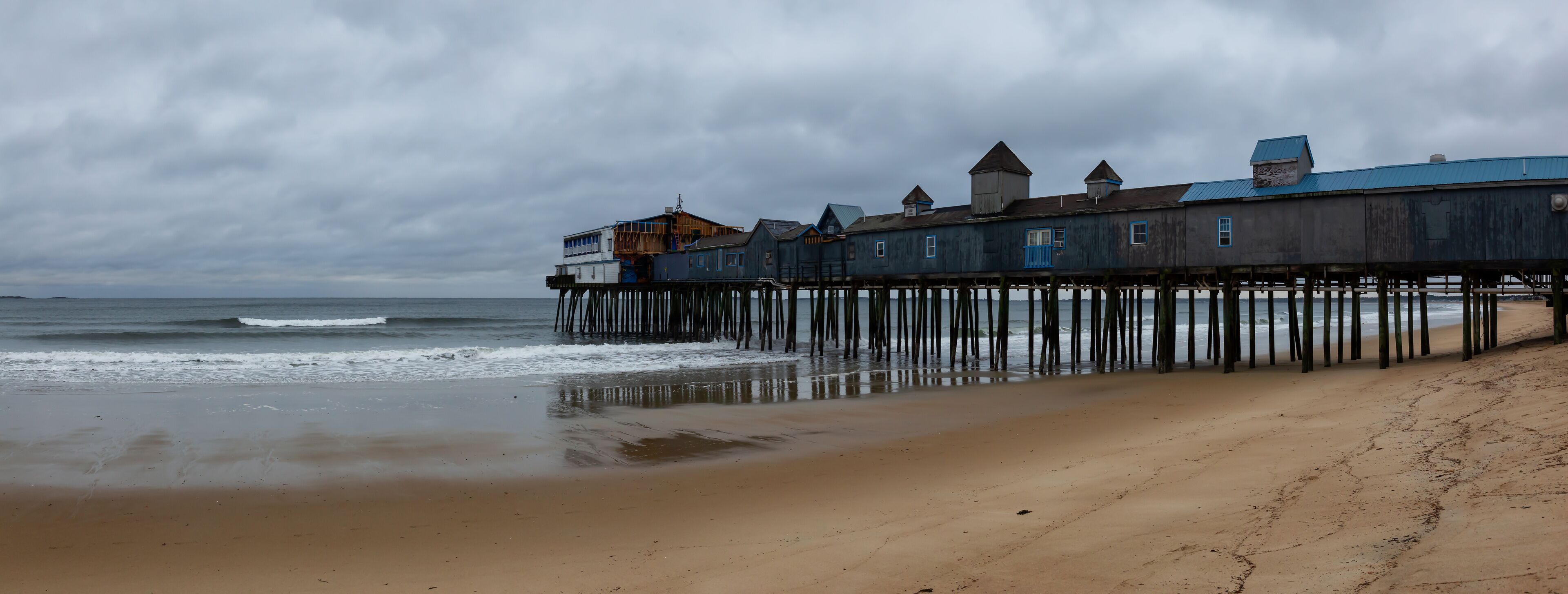 Panoramic view of a Wooden Pier on a sandy beach at the Atlantic Ocean during a cloudy sunrise. Taken in Old Orchard Beach, Maine, United States.