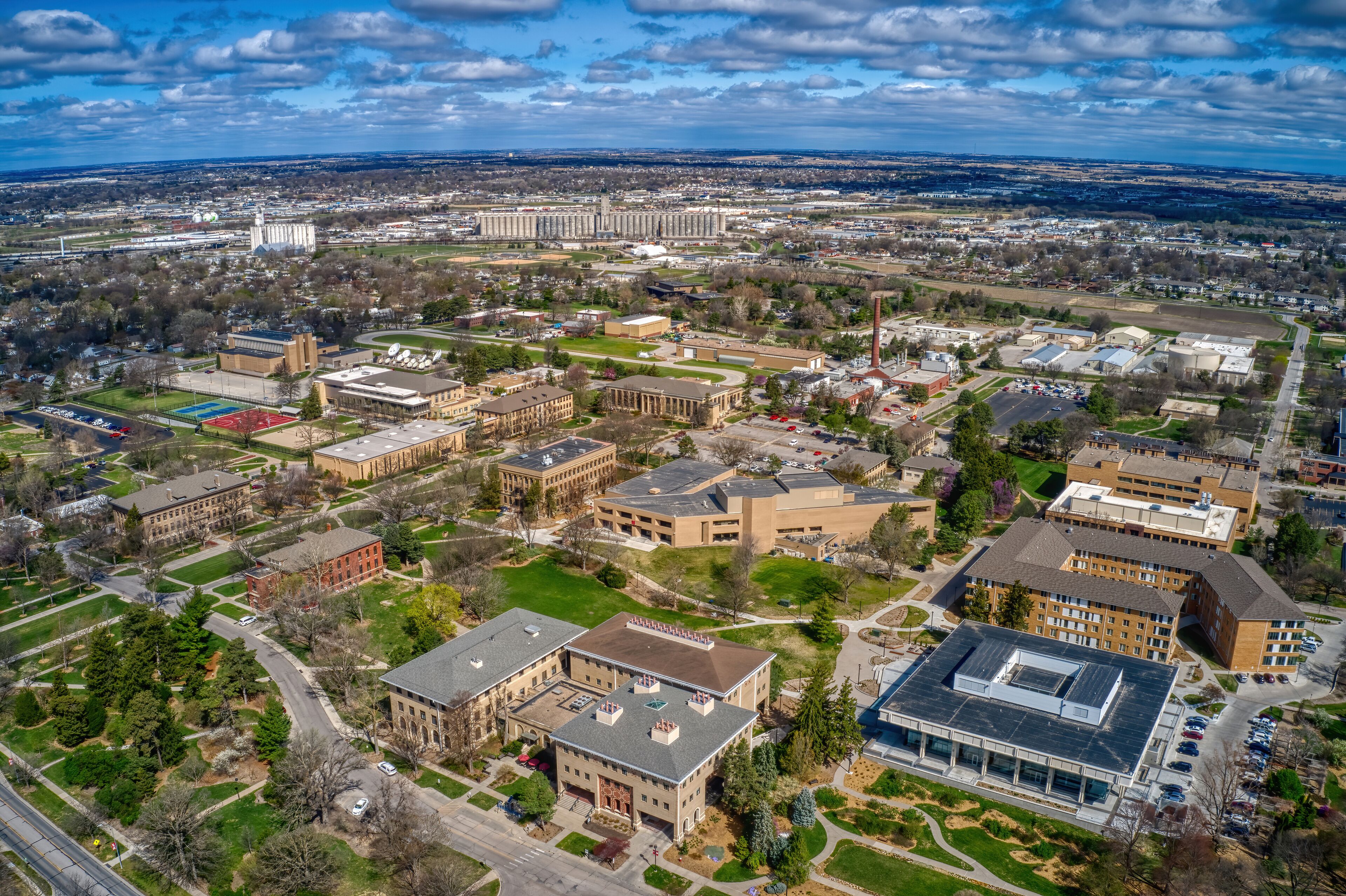 An aerial view of a university campus with multiple buildings, green spaces, and a distant city skyline.