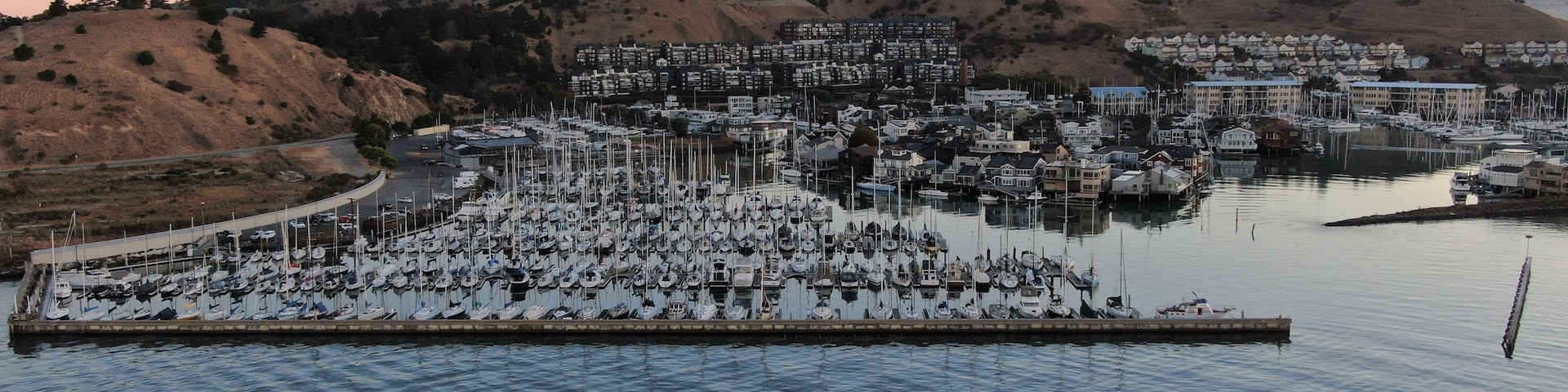 Uma marina com vários barcos atracados, rodeada por colinas e edifícios residenciais.