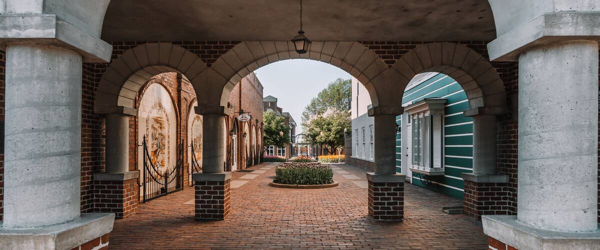 An arched brick corridor with a central garden and a green building in the background.