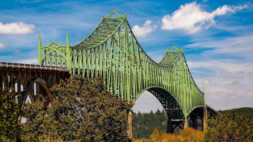 A green metal bridge with a unique arch design, surrounded by trees and hills.