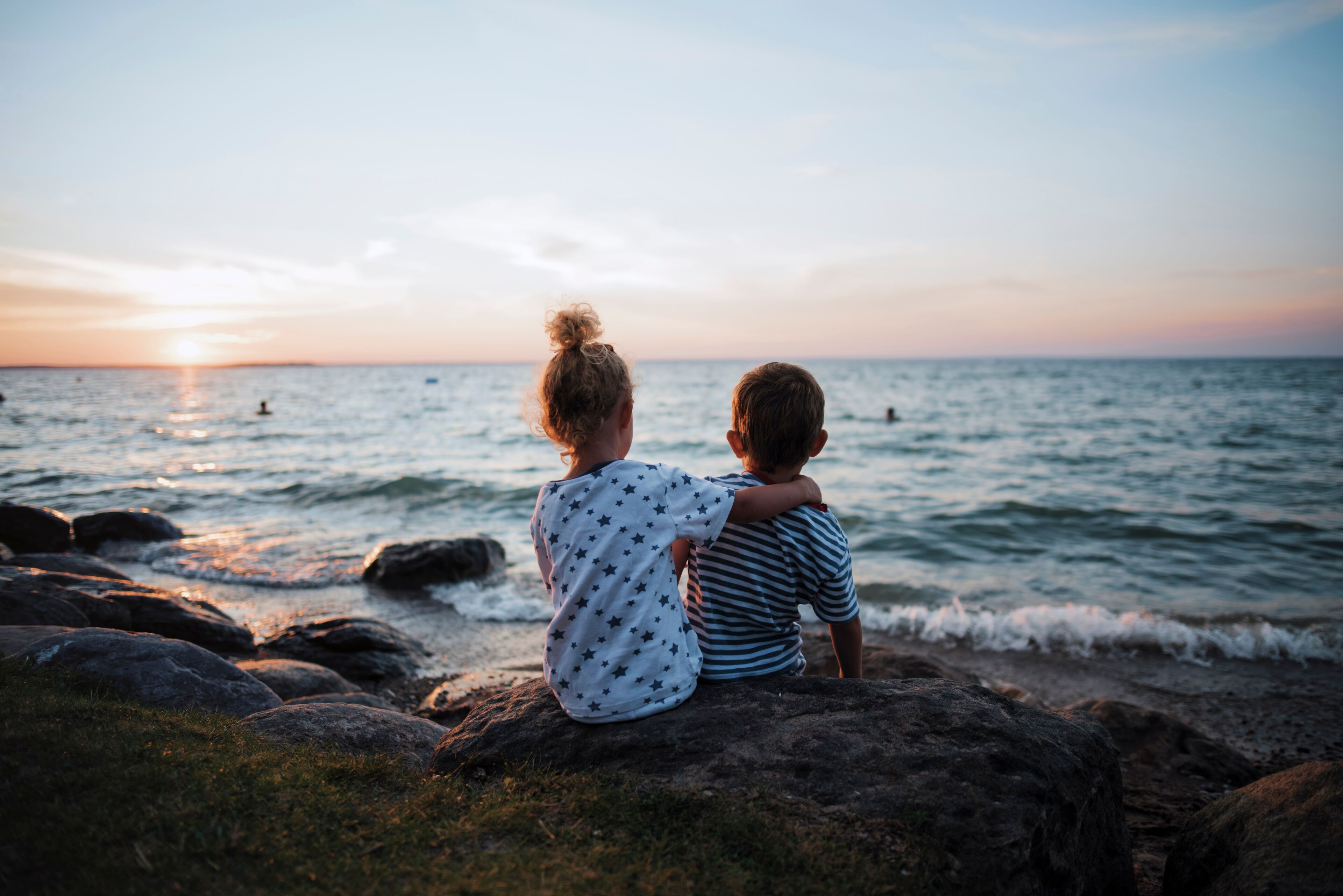 Rear view of siblings sitting on rocks by Lake Simcoe against sky during sunset