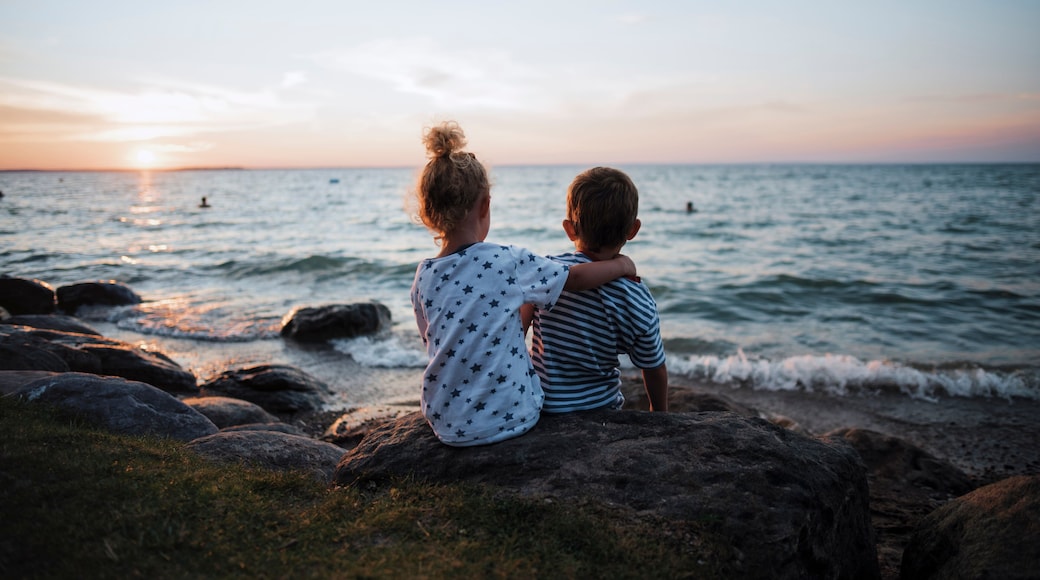 Rear view of siblings sitting on rocks by Lake Simcoe against sky during sunset