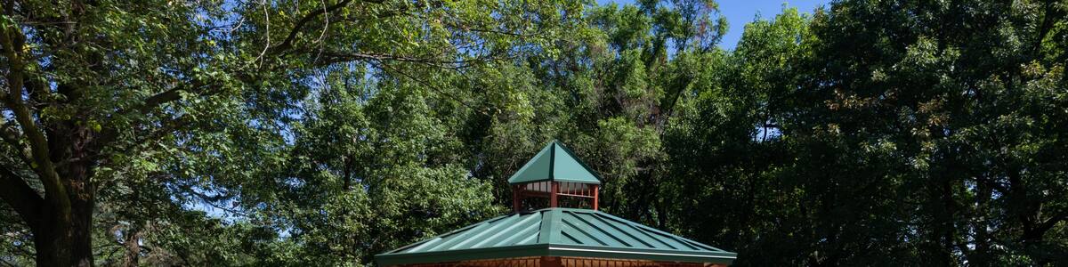 A gazebo with a green roof and brown pillars, surrounded by a small garden with shrubs and a bench.