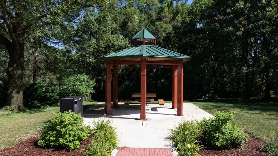 A gazebo with a green roof and brown pillars, surrounded by a small garden with shrubs and a bench.