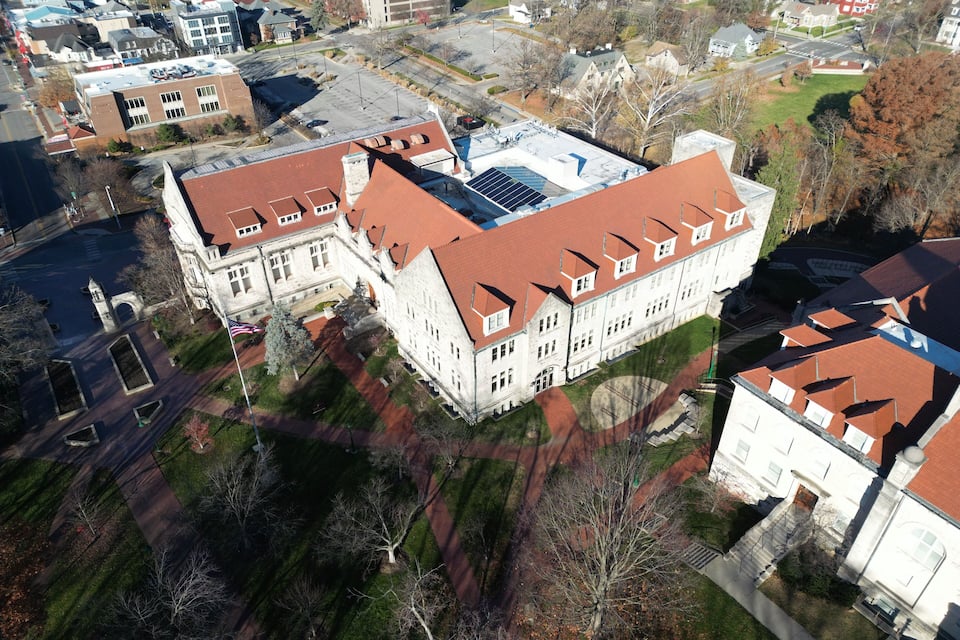 An aerial view of a large, historic building with a red roof, surrounded by trees and other buildings.