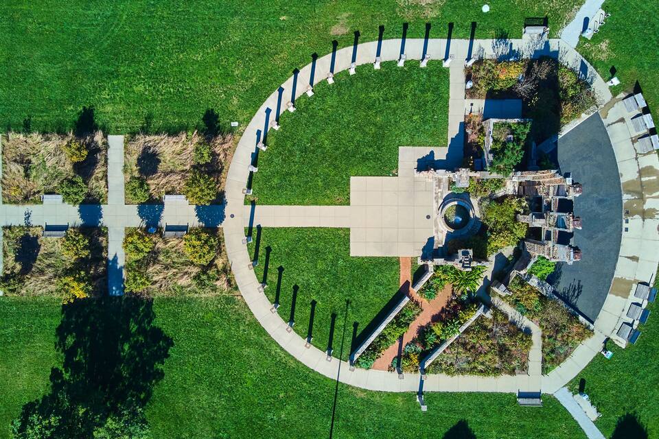 An aerial view of a park with a circular walkway, benches, trees, and a central monument.