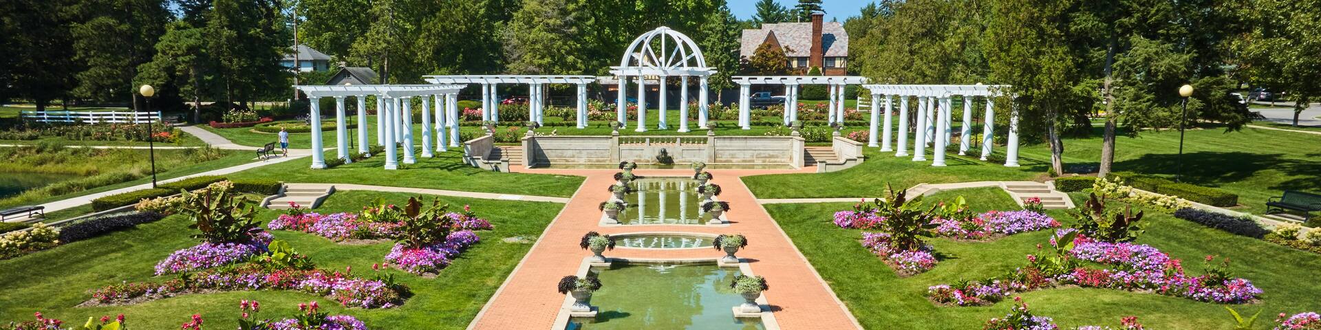 A formal garden with a central water feature, symmetrical flower beds, and white pergolas.