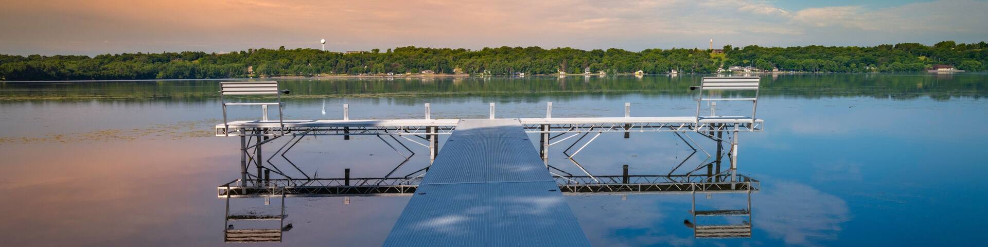 A calm lake with a dock extending into the water, surrounded by trees and a clear sky.