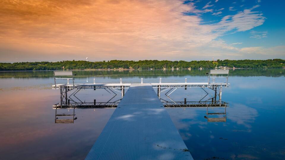 A calm lake with a dock extending into the water, surrounded by trees and a clear sky.