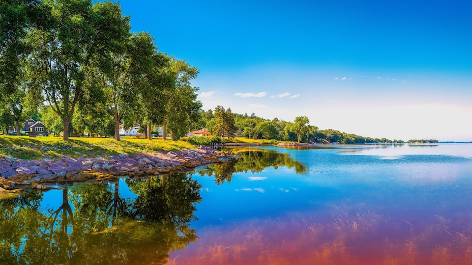A calm lake with reflections of trees and houses, surrounded by greenery and a clear sky.