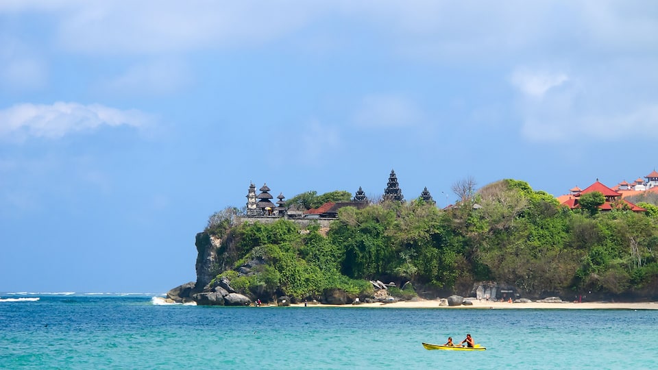 A coastal landscape with a temple on a cliff, a sandy beach, and a person kayaking.
