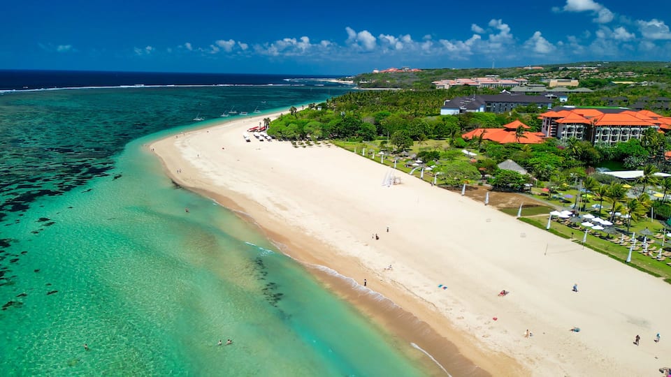 A coastal beach with clear turquoise waters, a sandy shore, and a resort area with buildings and umbrellas.
