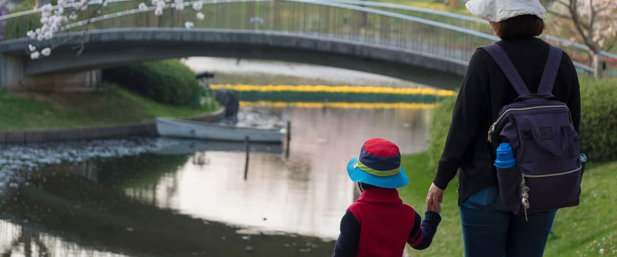 A woman and child walking on a bridge over a body of water, with cherry blossoms in the background.