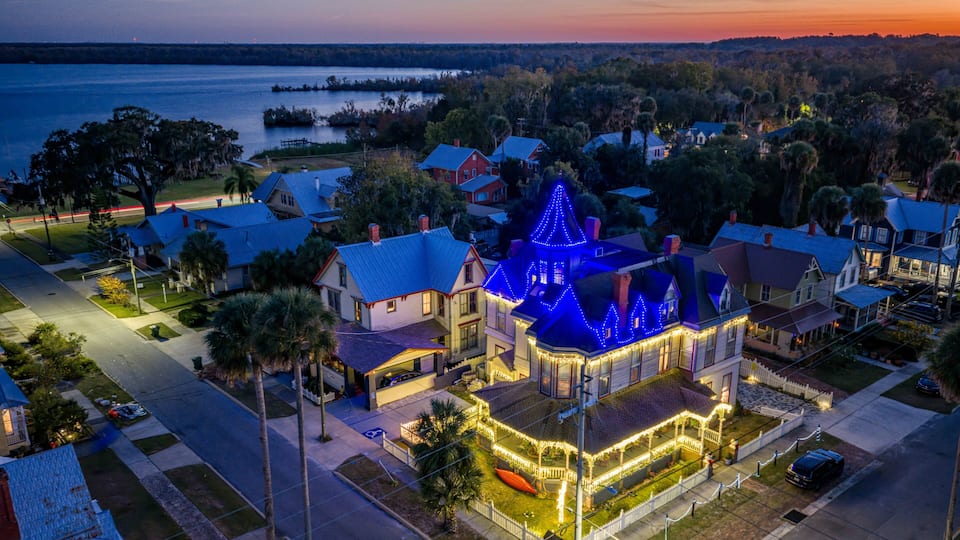 A residential area with houses, a prominent house with blue and white lights, and a body of water in the background.