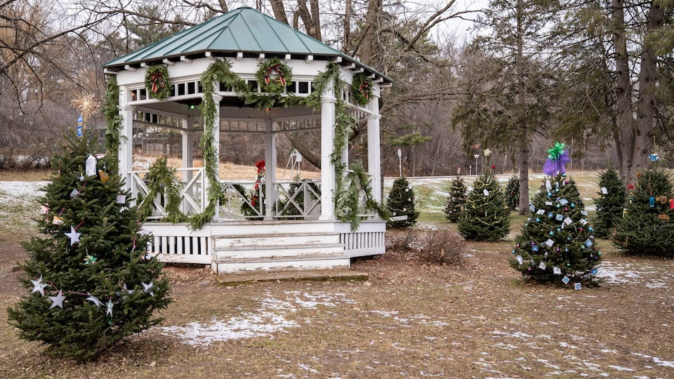 A gazebo decorated with Christmas wreaths and garlands, surrounded by small Christmas trees and snow on the ground.