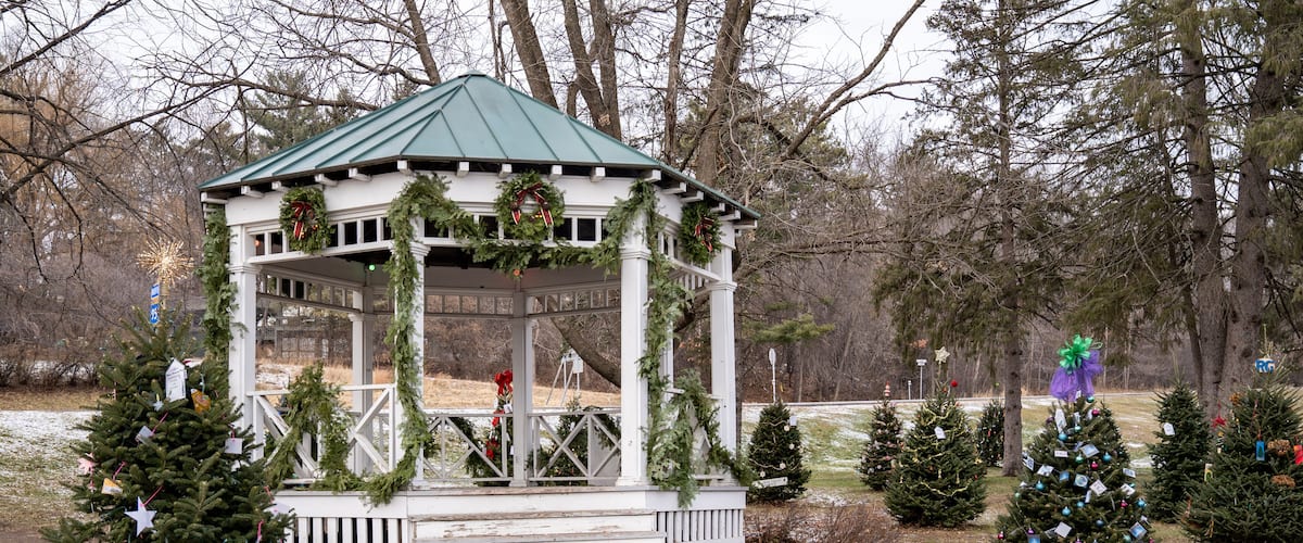 A gazebo decorated with Christmas wreaths and garlands, surrounded by small Christmas trees and snow on the ground.