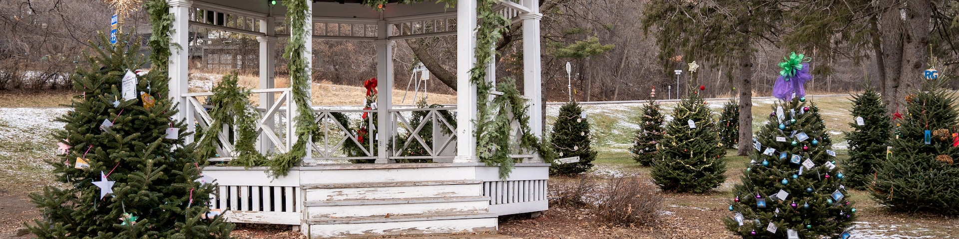 A gazebo decorated with Christmas wreaths and garlands, surrounded by small Christmas trees and snow on the ground.
