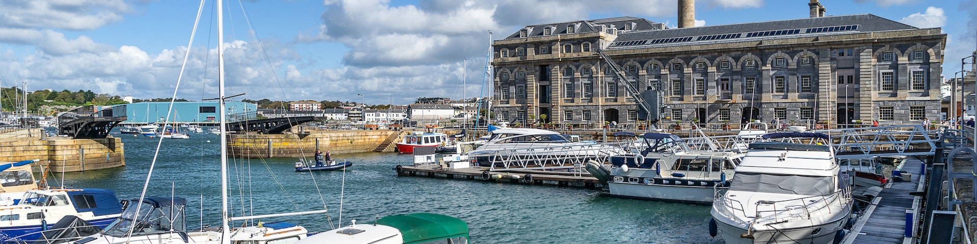 Um porto com vários barcos atracados, um edifício histórico imponente com uma torre destacada, e um céu azul límpido com algumas nuvens.