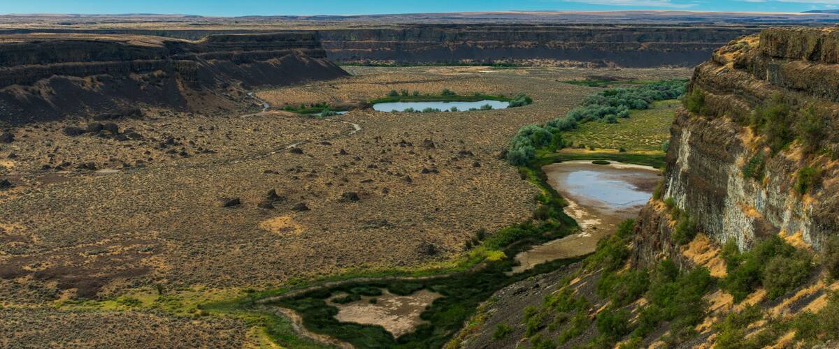 Een uitgestrekte kloof met een rivier die erdoorheen kronkelt, omringd door schaarse begroeiing en rotsachtig terrein.