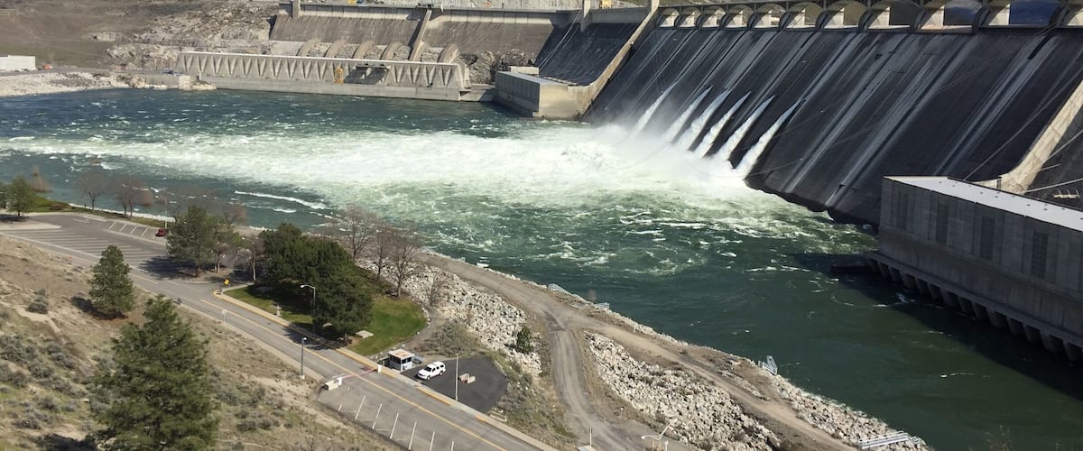 A dam with water cascading over its side, a road with a white vehicle, and a mountainous landscape in the background.