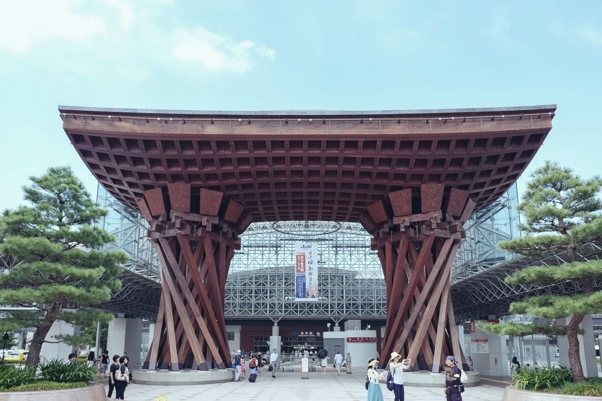 A large, open entrance with a metal roof and wooden beams, surrounded by trees and a glass structure in the background.