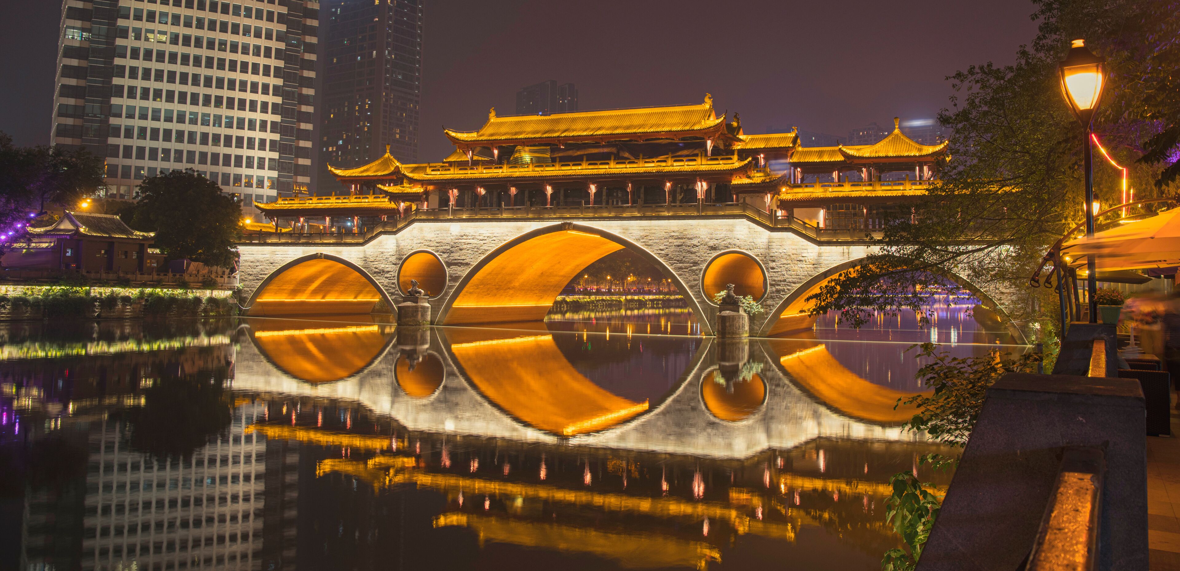 Un puente chino tradicional con techos dorados que se reflejan en el agua, rodeado de edificios modernos.