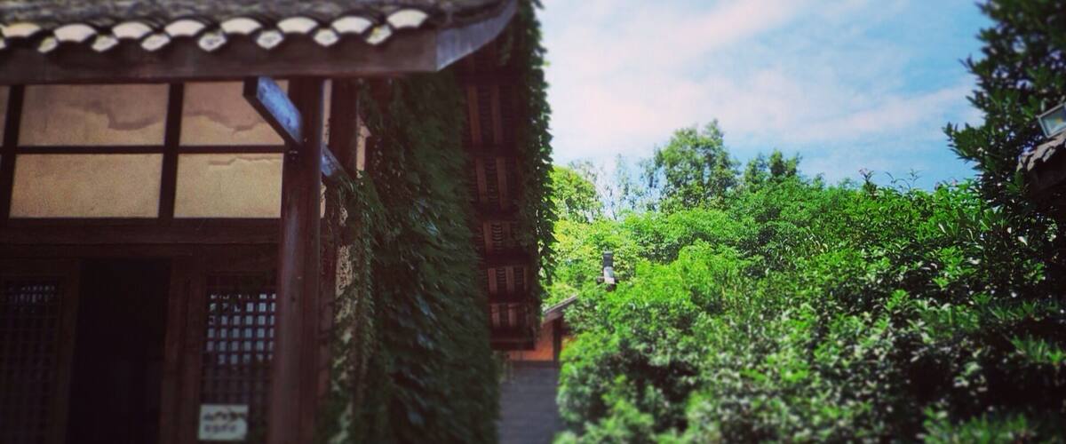 A traditional Japanese building with a tiled roof and wooden structure, surrounded by lush greenery and a clear sky.