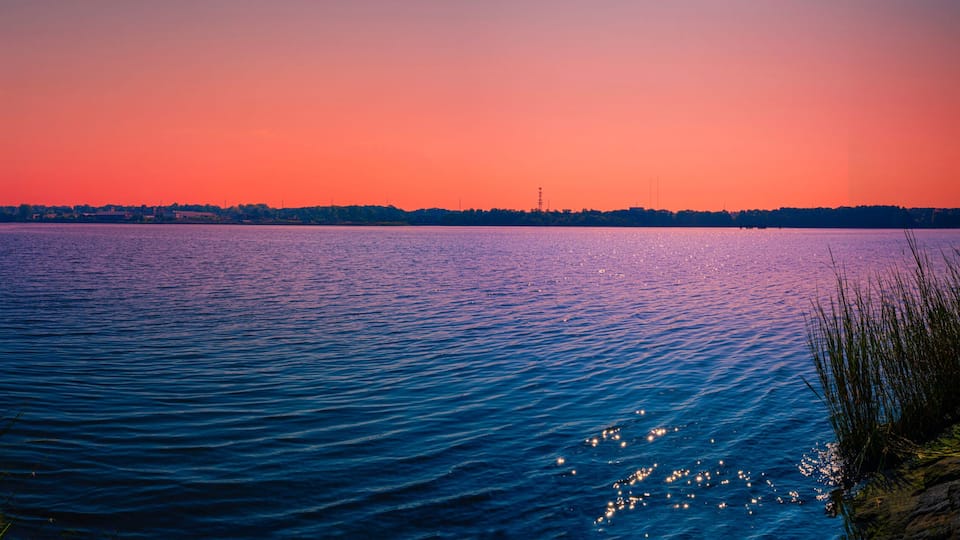 A calm lake at sunset with a forested shoreline and a distant city skyline.