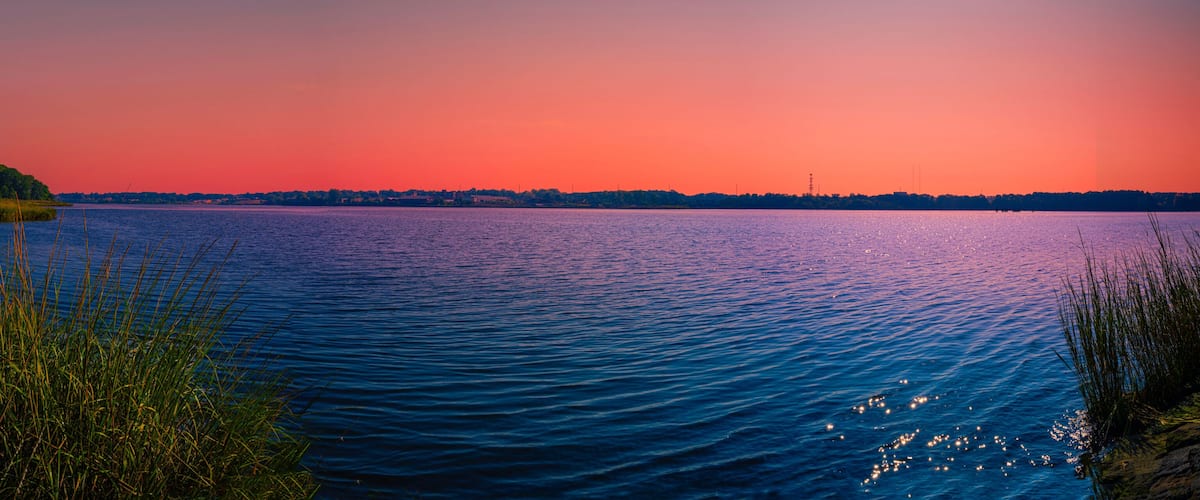 A calm lake at sunset with a forested shoreline and a distant city skyline.