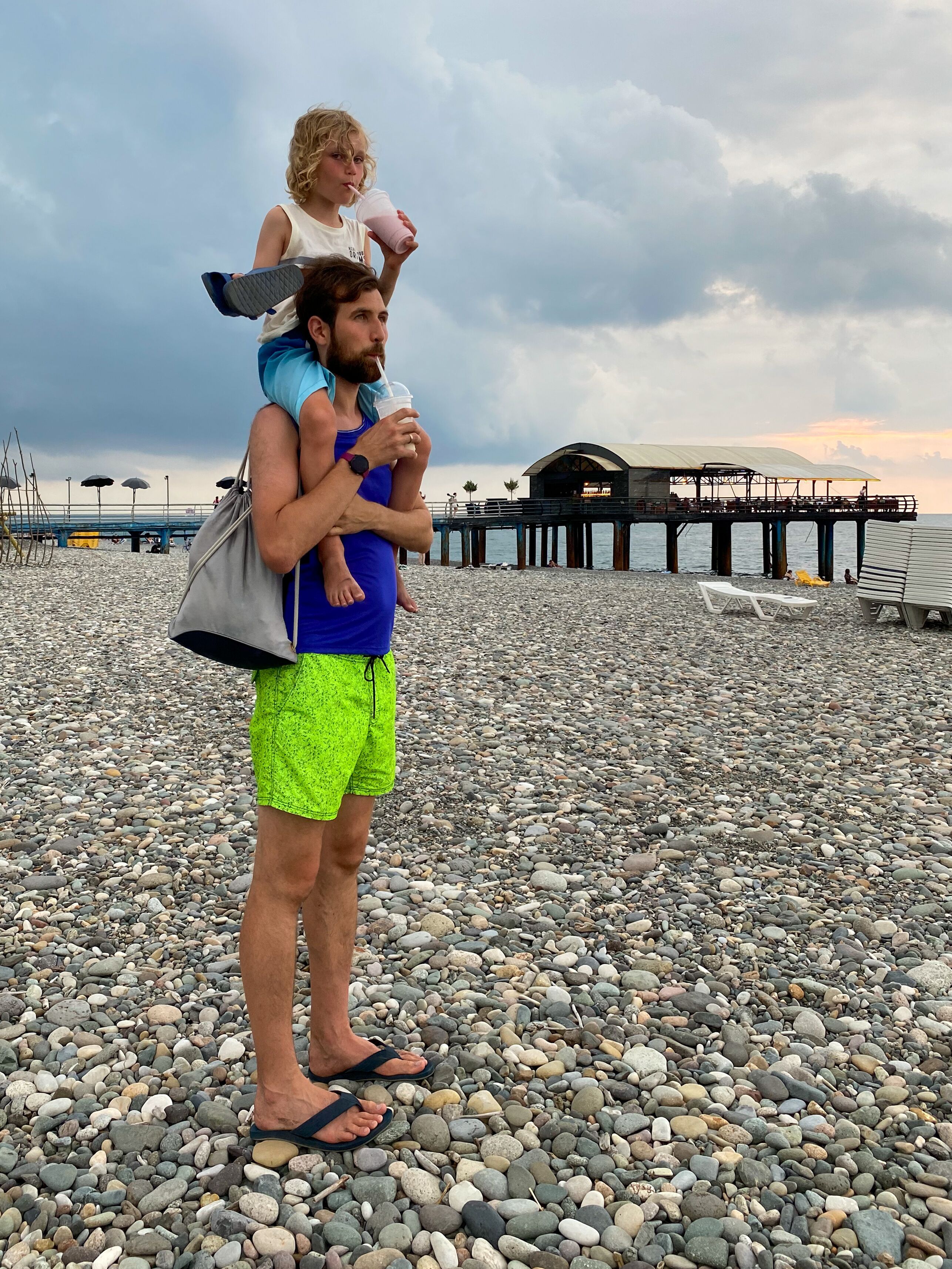Father Carrying Son On Shoulders Drinking Milkshakes On Pebble Beach Under Dramatic Storm Sky During Summer Walk